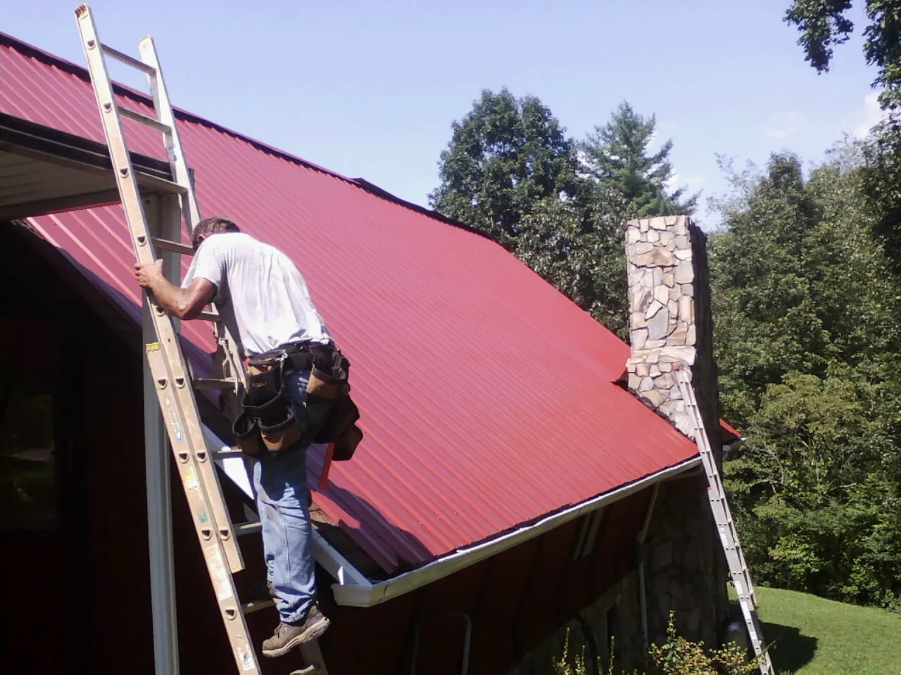 Roofer on a ladder beside a red metal roof and stone chimney — Soffit & Fascia Repair by Roofing Brothers in Caledonia